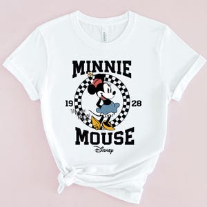May include: White t-shirt with a black and white chequered circle graphic featuring Minnie Mouse in a red dress and yellow shoes. The text "Minnie Mouse" is written in black letters above and below the circle. The text "1928" is written in black letters on either side of the circle. The text "Disney" is written in black letters below the graphic.