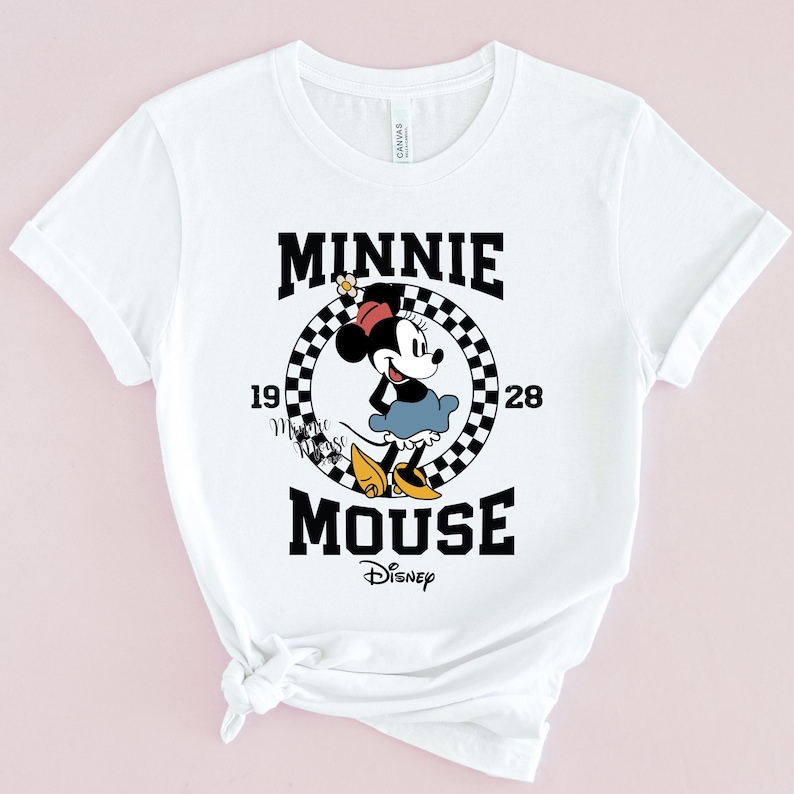 May include: White t-shirt with a black and white chequered circle graphic featuring Minnie Mouse in a red dress and yellow shoes. The text "Minnie Mouse" is written in black letters above and below the circle. The text "1928" is written in black letters on either side of the circle. The text "Disney" is written in black letters below the graphic.