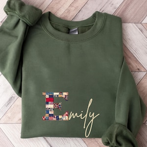Peut inclure: Sweat-shirt vert olive avec un 'E' en patchwork et le prénom 'Emily' en cursive. Le patchwork présente des motifs de tissus rouges, bleus et blancs. Le sweat-shirt a un col rond et des manches longues.