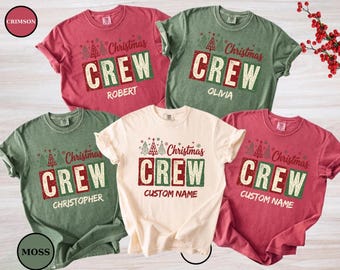 May include: Five short-sleeved t-shirts in crimson, moss green, and cream colors. Each shirt features the text "Christmas CREW" with stylized Christmas trees above and a personalized name printed below in a distressed, block-letter font style.