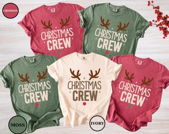 May include: A collection of five short-sleeved t-shirts in crimson, moss, and ivory. Each shirt features a reindeer antler design above the words "CHRISTMAS CREW" in a festive font. A red dot is below the text.