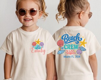 May include: An off-white short-sleeve t-shirt featuring a front graphic of a starfish, seashells, and the name Emily in pink script. The back displays the text Beach Crew, Miami FL, 2026, surrounded by colorful starfish, seashells, and coral.