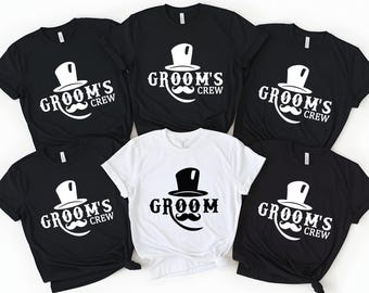 May include: A collection of black and white t-shirts. The shirts feature a top hat and mustache graphic with the text "Groom's Crew" in white. One shirt is white with a similar design and the word "Groom".