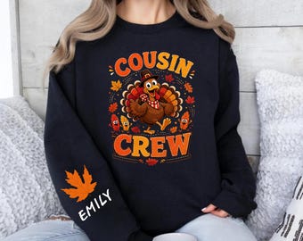 Może przedstawiać: Czarna bluza z długim rękawem z grafiką indyka w kapeluszu pielgrzyma i w kratkę szaliku, otoczona jesiennymi liśćmi i napisem COUSIN CREW. Na lewym rękawie znajduje się pomarańczowy liść klonu oraz biały napis z imieniem EMILY.