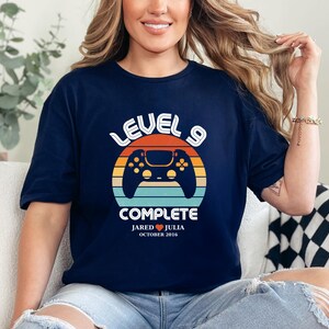 May include: A navy blue t-shirt with a retro-style graphic of a video game controller and the text "Level 9 Complete". The graphic also includes the text "Jared ♥ Julia October 2016".