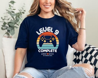 May include: A navy blue t-shirt with a retro-style graphic of a video game controller and the text "Level 9 Complete". The graphic also includes the text "Jared ♥ Julia October 2016".