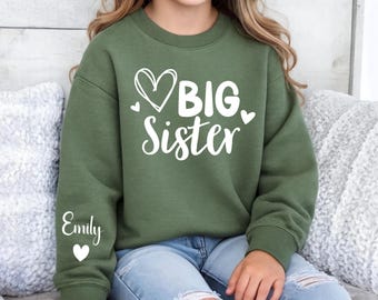 May include: An olive green sweatshirt with the words "BIG Sister" in white script and a heart design. The name "Emily" is printed on the left sleeve, also in white. The sweatshirt is worn by a child.