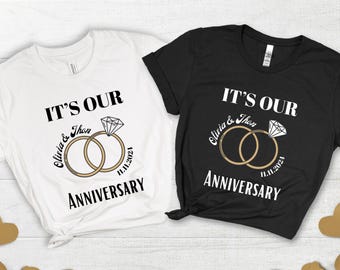 May include: Two t-shirts, one white and one black, with the text "IT'S OUR ANNIVERSARY" and the names "Olivia & Jhon" and the date "11.11.2021". The design includes two rings and a diamond.