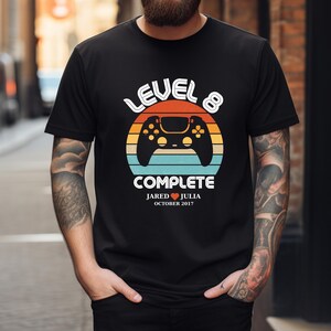 May include: Black t-shirt with a retro-style video game controller graphic. The text "Level 8 Complete" is above the controller, and the text "Jared ♥ Julia October 2017" is below the controller.
