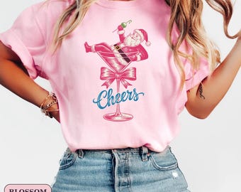 May include: Pink t-shirt with a graphic of Santa Claus relaxing in a martini glass. The image includes a pink bow and the word "Cheers" in blue script. The shirt is a light pink color.