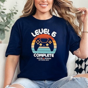 May include: A navy blue t-shirt with a retro-style graphic of a video game controller and the text "Level 6 Complete". The graphic also includes a sunset-like background with red, orange, and yellow colors. The text "David & Susan October 2019" is printed below the graphic.