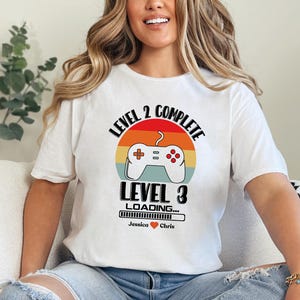 May include: White t-shirt with a retro-style video game controller graphic. The text reads "Level 2 Complete Level 3 Loading... Jessica Chris".