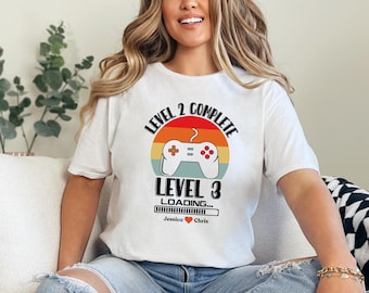 May include: White t-shirt with a retro-style video game controller graphic. The text reads "Level 2 Complete Level 3 Loading... Jessica Chris".
