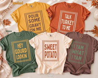 May include: Five short-sleeve crew neck t-shirts in mustard, rust, moss green, ivory, and espresso brown. Each shirt features a rectangular frame of vertical lines with text: POUR SOME GRAVY ON ME, TALK TURKEY TO ME, HEY GOURD LOOKIN', SWEET POTATO, and I YAM WHAT I YAM.