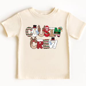 May include: A cream-colored t-shirt featuring the words "COUSIN CREW" in a festive, colorful design. The letters are decorated with Christmas-themed elements like snowmen, reindeer, and elves, making it a perfect holiday apparel item.