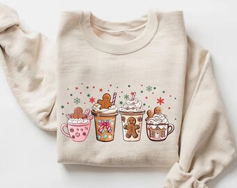 May include: Beige sweatshirt featuring a festive design of four holiday-themed drinks. Each drink is topped with whipped cream and a gingerbread man. The design includes red, green, and white decorative elements.
