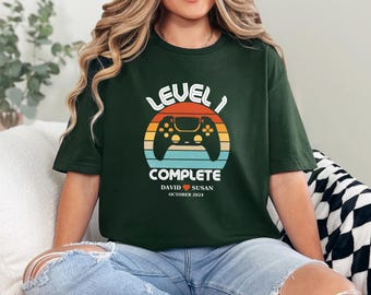 May include: Green t-shirt with a retro-style video game controller graphic and the text "Level 1 Complete" with the names "David" and "Susan" and the date "October 2024".