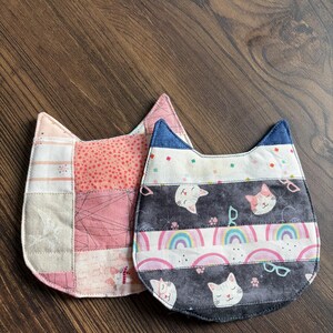 May include: Two cat-shaped fabric coasters. One is pink with various patterns, the other has a blue top and features cat faces, rainbows, and glasses on a dark gray background. The coasters are on a wooden surface.