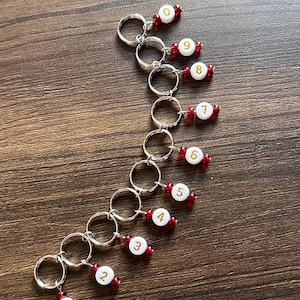 May include: Ten silver stitch markers with red beads and white numbers from 0 to 9. The markers are arranged in a curved line on a wooden surface.