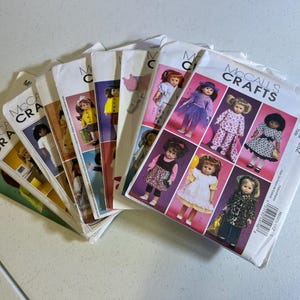 May include: A stack of McCall's Crafts sewing patterns for dolls. The patterns feature images of dolls in various outfits, including dresses, pajamas, and coats. The patterns are labeled with the text "McCall's Crafts" and the code "M6005".