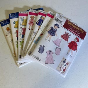May include: A stack of vintage Simplicity sewing patterns for baby doll clothes. The patterns feature illustrations of dresses, coats, and other garments. The patterns are in various sizes and styles, with the Simplicity logo visible.