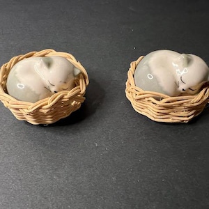 May include: Two ceramic sleeping cat figurines in woven baskets. The cats are light gray with closed eyes, curled up in a sleeping position. The baskets are made of light brown woven material.