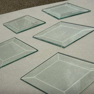 Op de afbeelding: Vijf heldere, ruitvormige decoratieve spiegels van glas met afgeschuinde randen. De spiegels zijn gerangschikt op een lichtgrijze ondergrond, wat hun reflecterende kwaliteit en geometrische ontwerp benadrukt. De spiegels variëren in grootte.