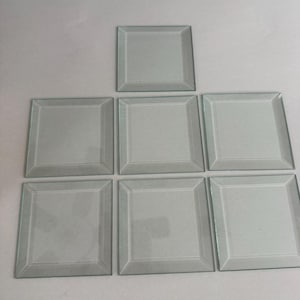 Könnte beinhalten: Sieben klare quadratische Untersetzer aus Glas mit abgeschrägter Kante sind auf einer weißen Oberfläche angeordnet. Die Untersetzer sind hellgrün und haben ein schlichtes, elegantes Design. Sie eignen sich für Getränke.