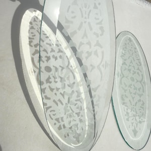 May include: Two clear, oval-shaped glass mirrors with etched floral patterns. The mirrors are positioned at an angle, casting a shadow on a white surface. The design is visible on the glass.