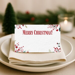 Puede incluir: Una tarjeta de mesa blanca con el texto "MERRY CHRISTMAS!" en rojo. Está decorada con ilustraciones de acuarela de bayas rojas y hojas verdes. La tarjeta está colocada sobre un plato blanco con una servilleta doblada.
