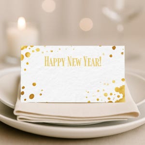 Może przedstawiać: Biała karta ze złotym napisem "HAPPY NEW YEAR!" umieszczona na białym talerzu z beżową serwetką. Złote plamy farby zdobią krawędzie karty, tworząc świąteczny wzór. W tle zapalona świeca.