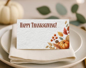 May include: A white place card with the text "HAPPY THANKSGIVING!" in brown. The card features a watercolor illustration of a pumpkin, berries, and fall leaves. The card is placed on a white napkin and plate.
