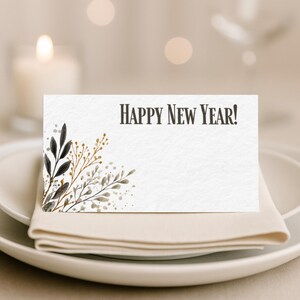 Peut inclure: Une carte de place blanche avec le texte "HAPPY NEW YEAR!" en marron foncé. La carte présente des accents botaniques de style aquarelle en noir, or et gris. La carte est posée sur une assiette blanche avec une serviette beige pliée.