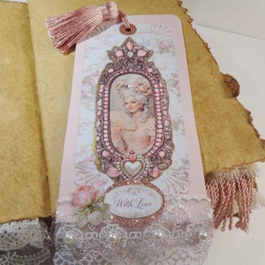 May include: A decorative bookmark featuring a portrait of a woman in a pink frame, adorned with a pink tassel. The bookmark has a floral design and the words "With Love". The bookmark is embellished with lace, pearls, and a pink rose.