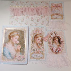 May include: A collection of vintage-style greeting cards and stationery. The items feature floral patterns, lace, and images of a woman smelling a rose. Colors include pastel pink, blue, and white. Some cards have the word "Love" or "Merci".
