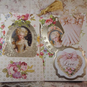 May include: A decorative paper craft with a floral design, featuring portraits in ornate frames, a heart-shaped embellishment with the word "Love", and a pair of lace gloves. A gold tassel hangs from the top.