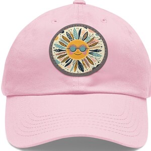 Peut inclure: Casquette de baseball rose clair avec un écusson circulaire. L'écusson représente un soleil souriant portant des lunettes de soleil, avec des détails jaunes, oranges et bleus, entouré de feuilles et de plumes. La casquette a une visière incurvée.