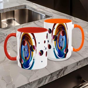 May include: Two white ceramic mugs with colorful abstract designs. One mug has a red interior and handle, the other has an orange interior and handle. The mugs are on a marble countertop in a kitchen setting.