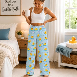 May include: Light blue pajama pants featuring a repeating pattern of yellow rubber ducks and colorful bubbles. The pants have a relaxed, wide-leg fit and an elastic waistband, showcasing a playful design with various duck poses and floating bubbles.