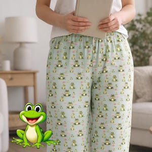May include: Light blue pajama trousers featuring a repeating pattern of cartoon frogs and small pink flowers. The trousers are made of a soft, lightweight fabric. The person in the image is holding a book.