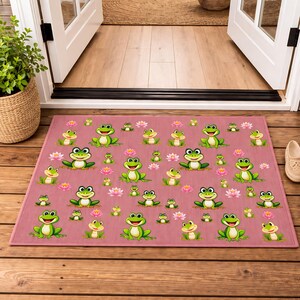 May include: A rectangular doormat with a pink background features a repeating pattern of cartoon frogs and pink water lilies. The frogs are green and white with smiling faces. The mat is placed in front of a white door.