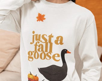 May include: White long-sleeved top with the text "just a fall goose" in gold. It features a black goose with orange feet and beak, and a small red wagon with pumpkins. An orange leaf is above the text, perfect for autumn.