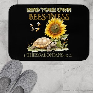 May include: Black bath mat with a yellow sunflower, a turtle, and bees. The text "Mind Your Own Bees-iness" and "1 Thessalonians 4:11" are printed on the mat.