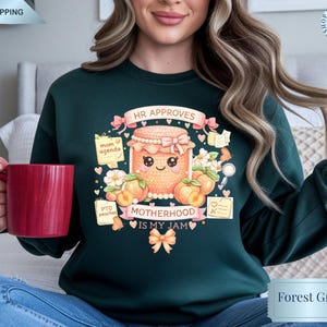 Può includere: Felpa verde foresta con una grafica colorata di un barattolo di marmellata sorridente, pesche e la scritta "MOTHERHOOD IS MY JAM". Il design include le parole "HR APPROVES", "mom agenda" e "PTO peaches".