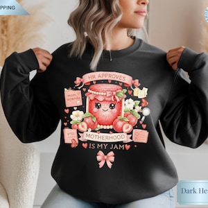Peut inclure: Sweat-shirt gris foncé avec un motif coloré. Le motif représente un pot de confiture souriant avec le texte "Motherhood is my jam". Le texte supplémentaire comprend "HR Approves", "Mom Agenda" et "PTO".