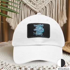 Può includere: Cappellino da baseball bianco con una toppa nera con un disegno di tartaruga blu e la scritta "TURTLE DAD 2024". Il cappellino ha una visiera curva e un classico design a sei pannelli. La toppa è rettangolare con un bordo cucito.
