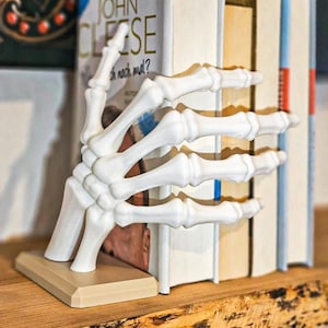 May include: A white, skeletal hand bookend holds several books on a wooden shelf. The bookend is made of a hard material and has a beige base. The books have various colors and titles, creating a unique decorative piece.