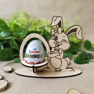 May include: Wooden Easter bunny egg holder with a chocolate egg inside. The bunny is holding its arms crossed and has a bow tie. The egg is a Kinder Surprise egg with the text "Kinder Überraschung" on it.