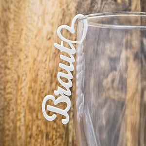 May include: A white, laser-cut drink charm with the word "Braut" in a cursive font, attached to a heart-shaped design. The charm is placed on the rim of a clear glass, with a wooden background.
