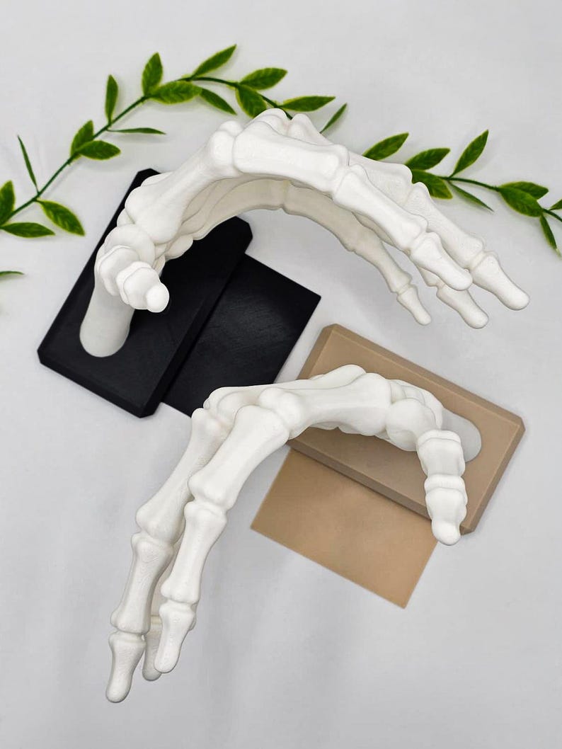 May include: Two white, skeletal hand sculptures, one on a black base and the other on a tan base. The hands are arched, with fingers extended. Green leafy vines are in the background.